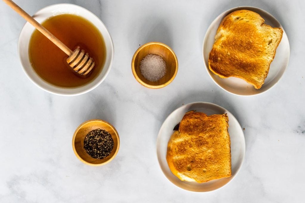 Ingredients on a counter including a bowl of honey, pinch bowls of sea salt and pepper and two slices of toast.