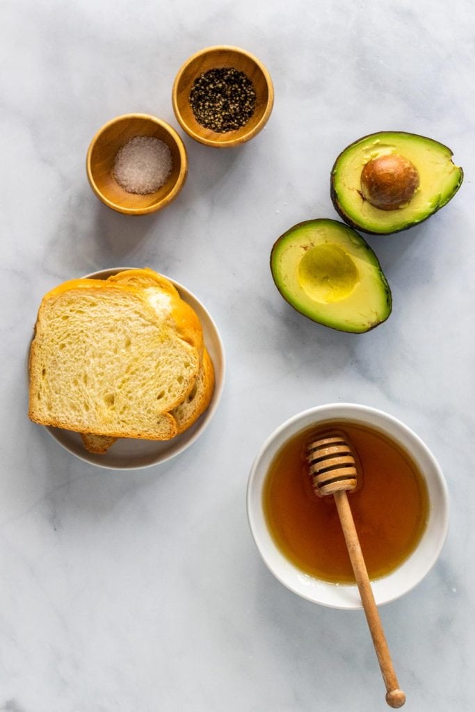 Ingredients spread out on the kitchen counter: avocado toast sliced in half, a bowl of honey, pinch bowls of salt and pepper, and two slices of bread.