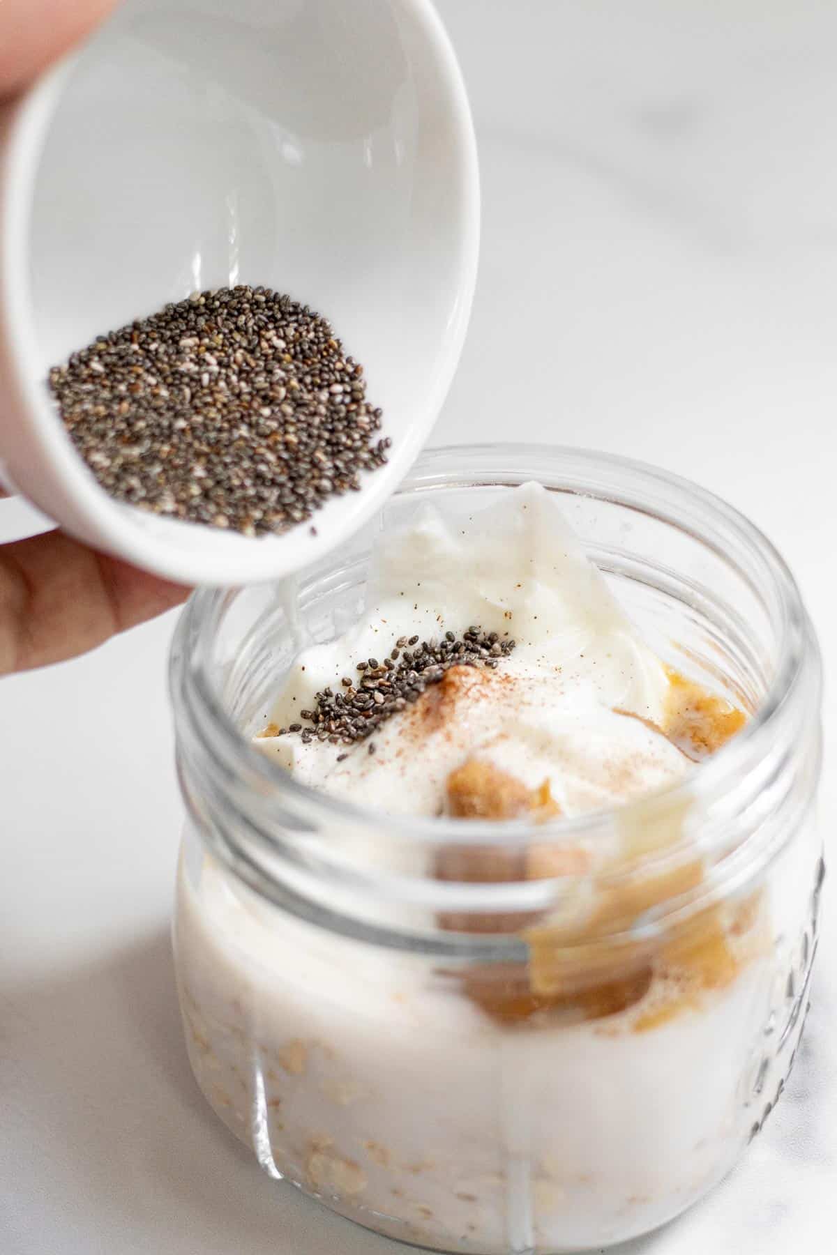 Pouring chia seeds into a 12 oz jar that contains the other ingredients for overnight oats with protein powder.