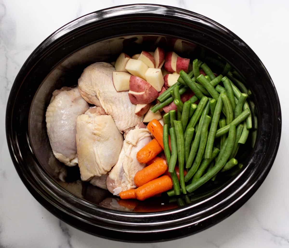 Raw chicken thighs, green beans, baby carrots, and cubed red potatoes in a slow cooker.