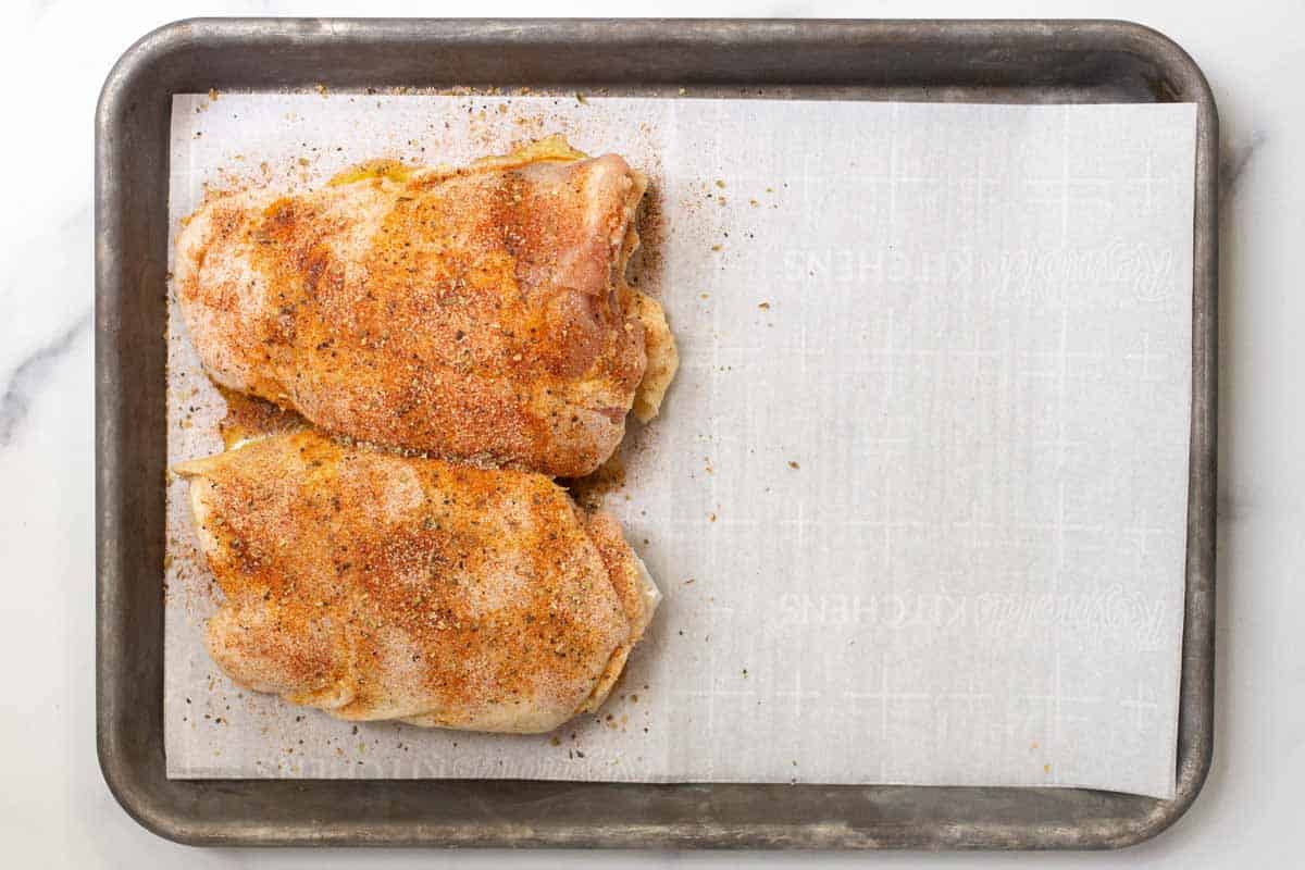 Seasoned raw chicken on one side of the parchment-lined rimmed baking sheet.
