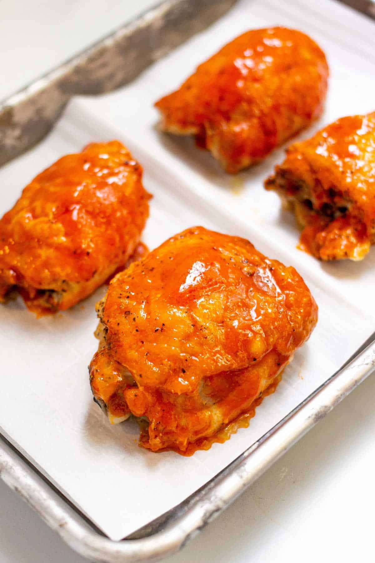 Four baked chicken thighs covered in buffalo sauce on a parchment lined baking sheet.