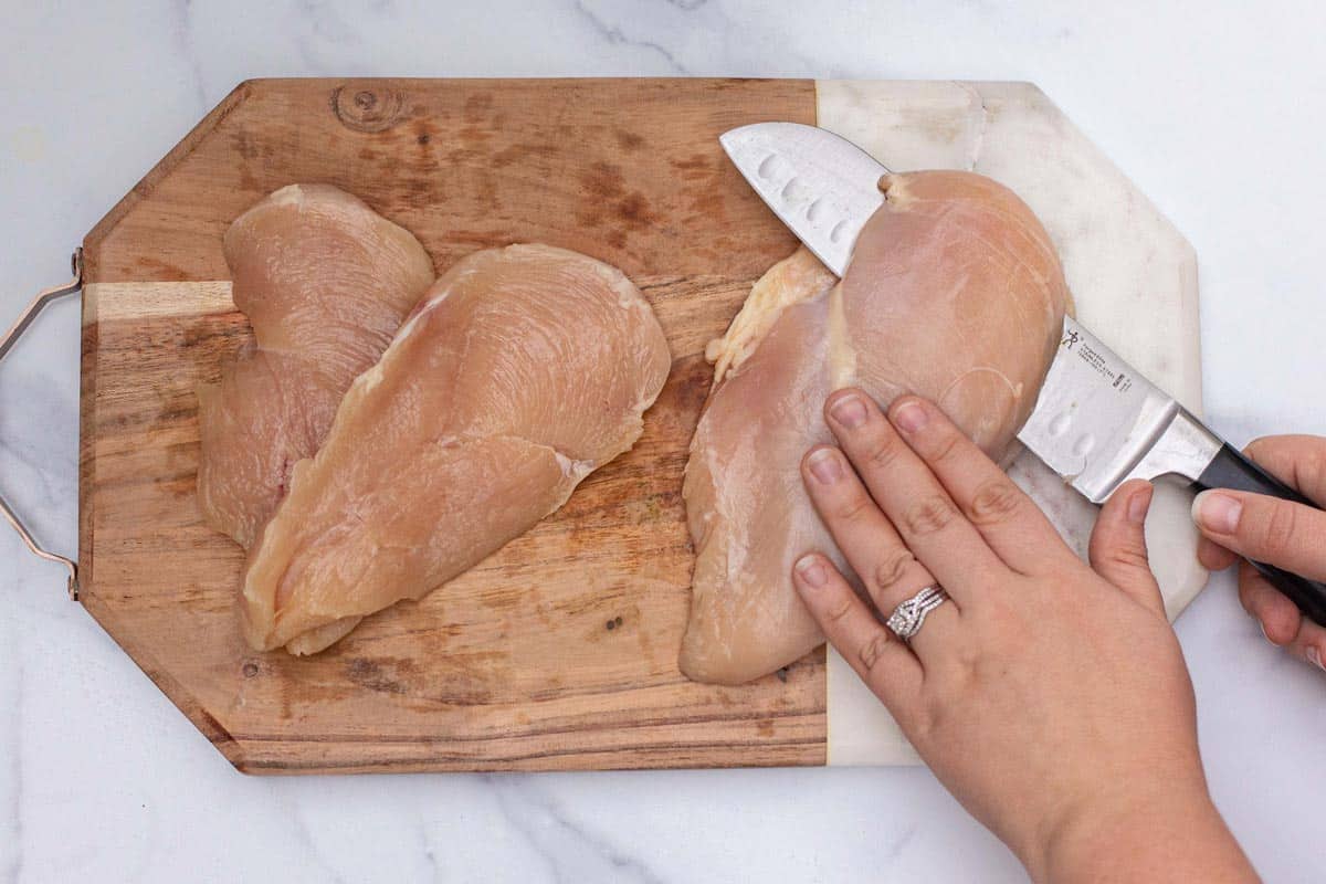 Cutting raw chicken breast in half horizontally with a knife while holding breast flat with the other hand.