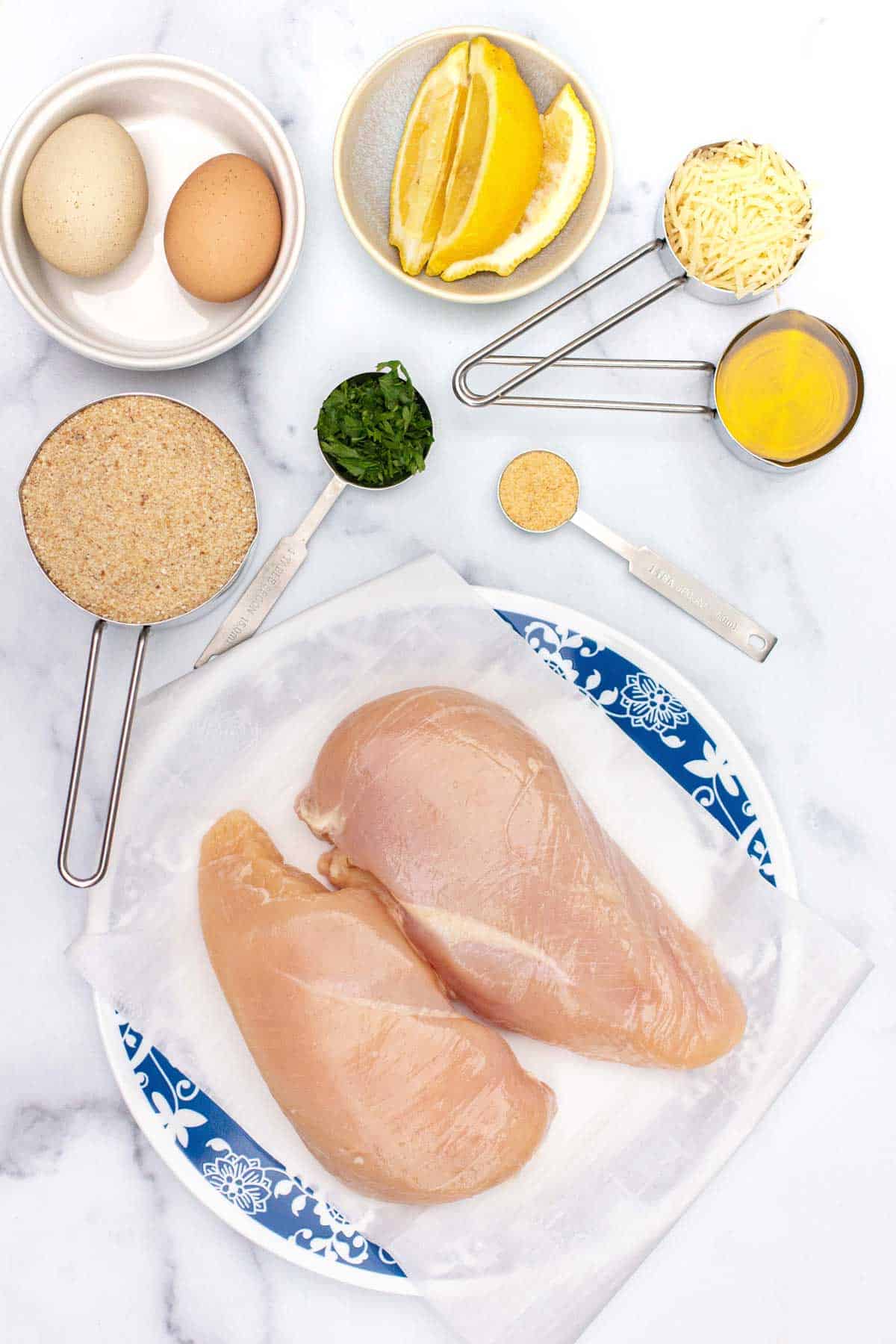 Italian chicken cutlet ingredients: raw chicken breasts, bread crumbs, parmesan, eggs, parsley, onion powder, olive oil, and lemon wedges.