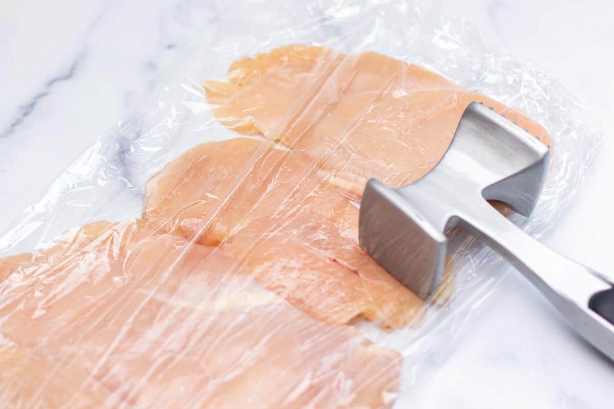 Raw chicken breast between two layers of plastic wrap pounded thin with meat mallet.