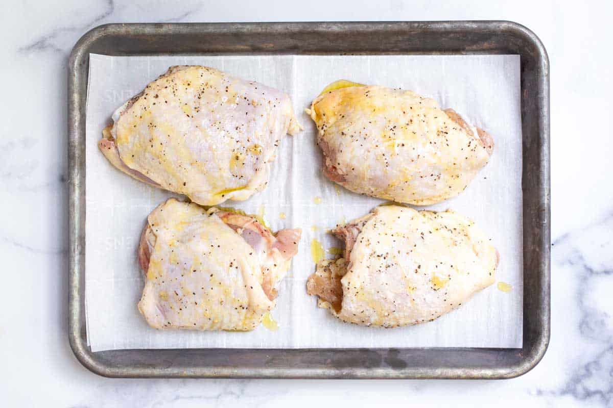 Four raw bone in skin on chicken thighs covered in seasoning and olive oil on a parchment lined baking sheet.