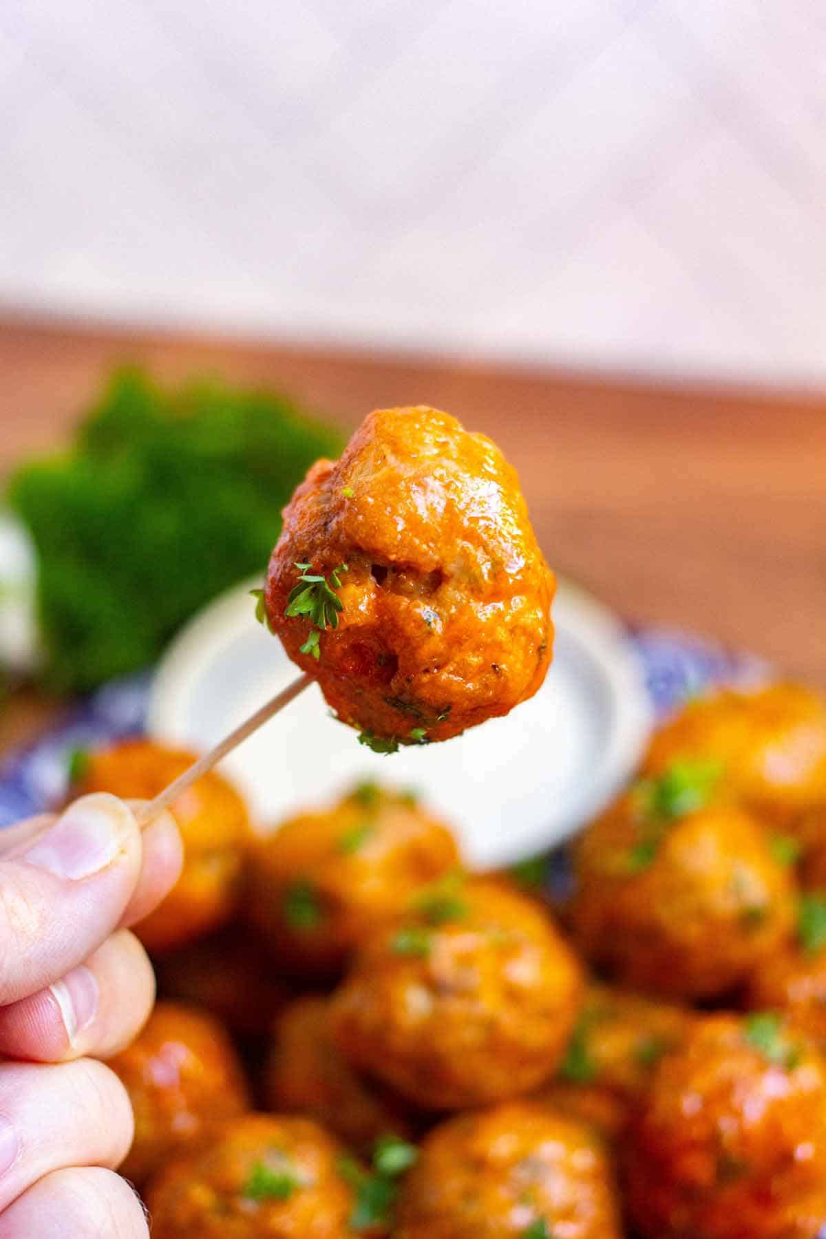 Buffalo turkey meatballs being held up on a toothpick. There is a plate of buffalo turkey meatballs in the background.