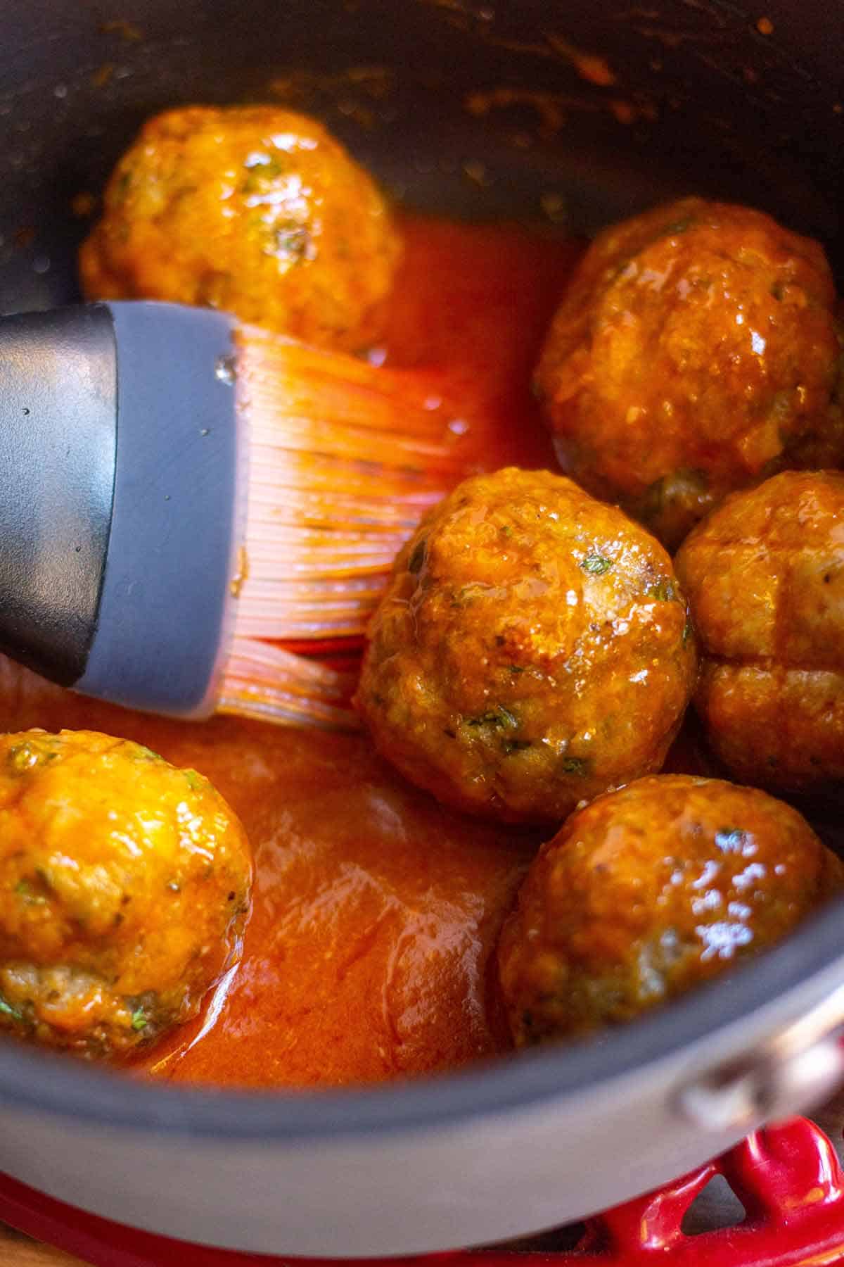 Cooked buffalo turkey meatballs in a saucepan that has buffalo sauce in it. A silicone basting brush is being used to coat the meatballs in sauce.