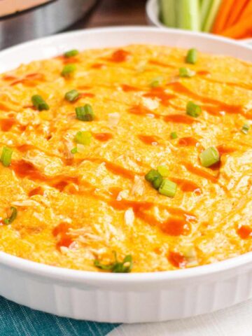 Close up of Buffalo chicken dip in a large 11 inch round baking dish. The dip is garnished with a drizzle of hot sauce and sliced green onions.