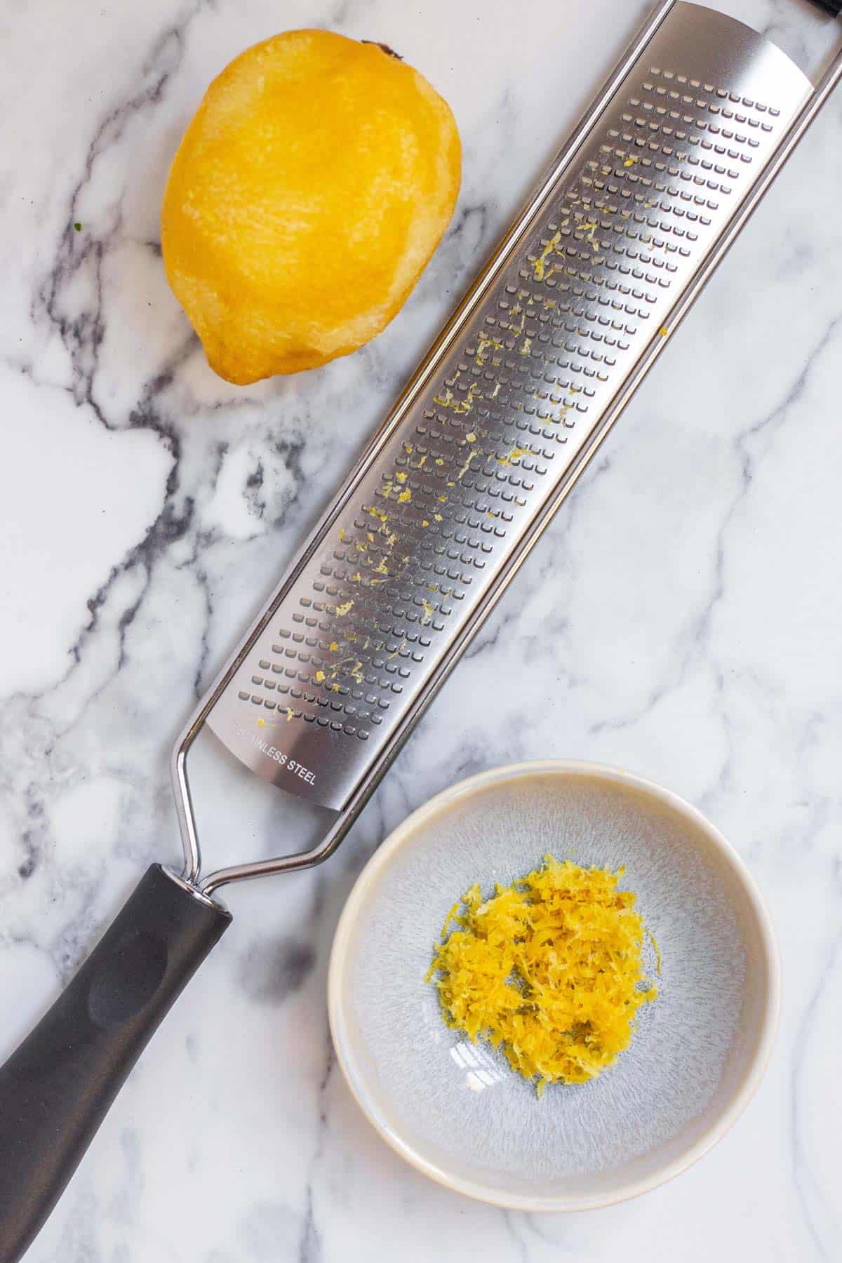 A small pinch bowl filled with fresh lemon zest next to a whole lemon and a microplane zester.