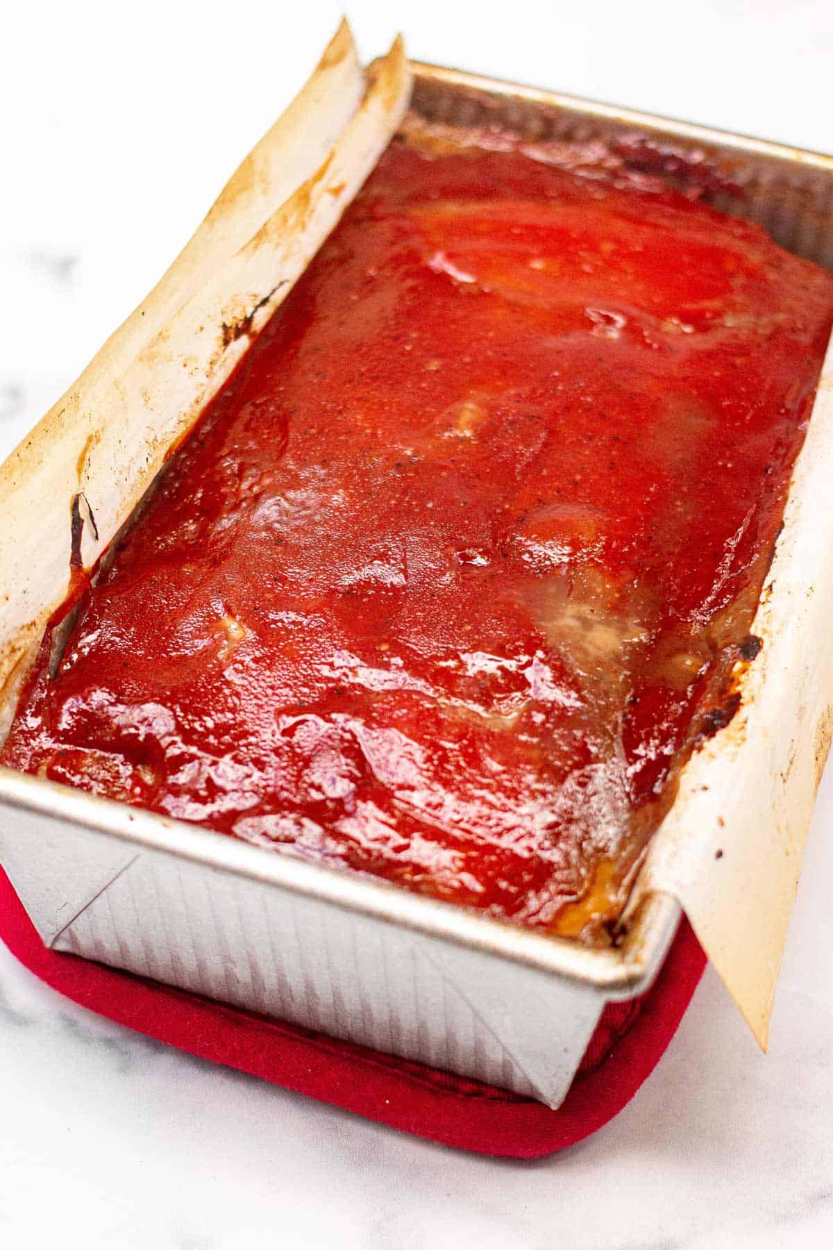 Baked meatloaf covered in ketchup glaze in a loaf pan sitting on a hot mitt. The parchment is yellowed from baking.
