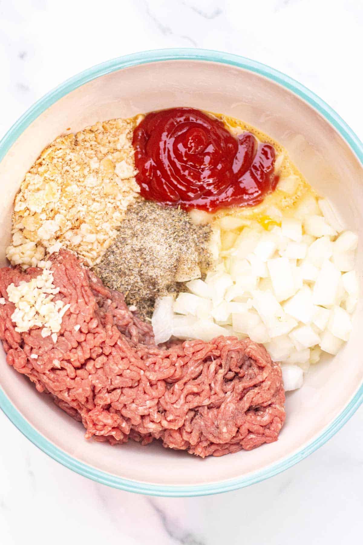 Meatloaf ingredients unmixed in a large bowl: lean ground beef, Ritz crackers crumbled, finely diced onion, minced garlic, beaten eggs, milk, ketchup, Italian seasoning, salt, ground black pepper, and garlic powder.