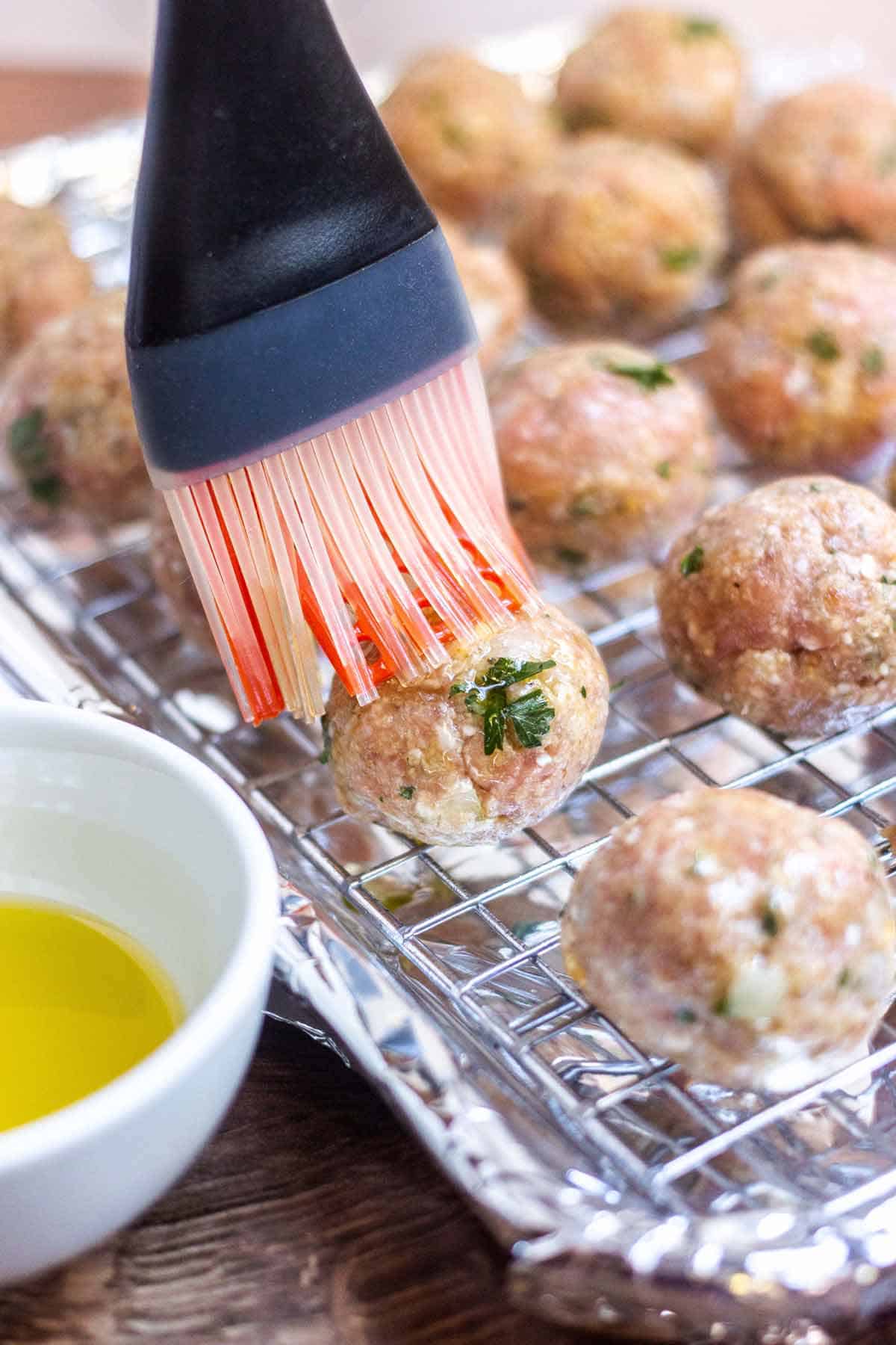 A silicone basting brush is being used to cover raw turkey meatballs with olive oil. The meatballs are on a wire rack on a baking tray that is covered in foil.