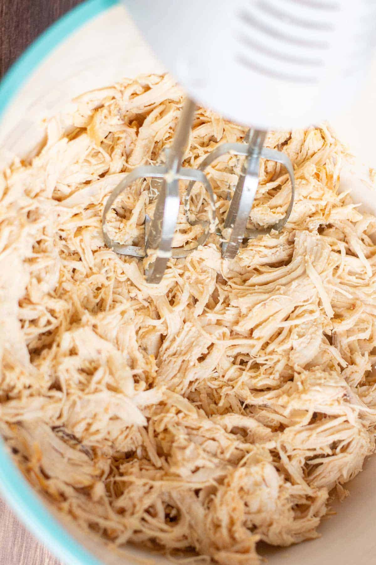 Large bowl of shredded chicken. A hand mixer is being used to shred the chicken.