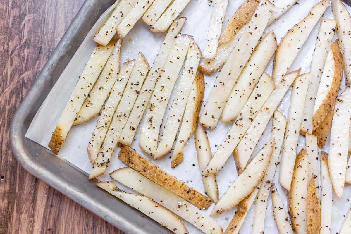 A sheet pan lined with parchment paper and covered with uncooked french fries coated in oil and black pepper.