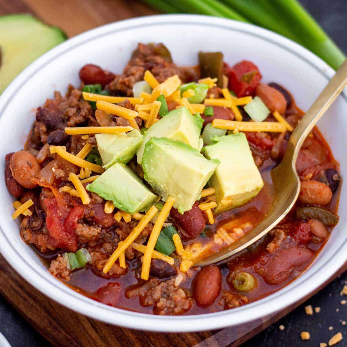 Small bowl of ground beef and sausage chili with shredded cheddar and diced avocado on top.