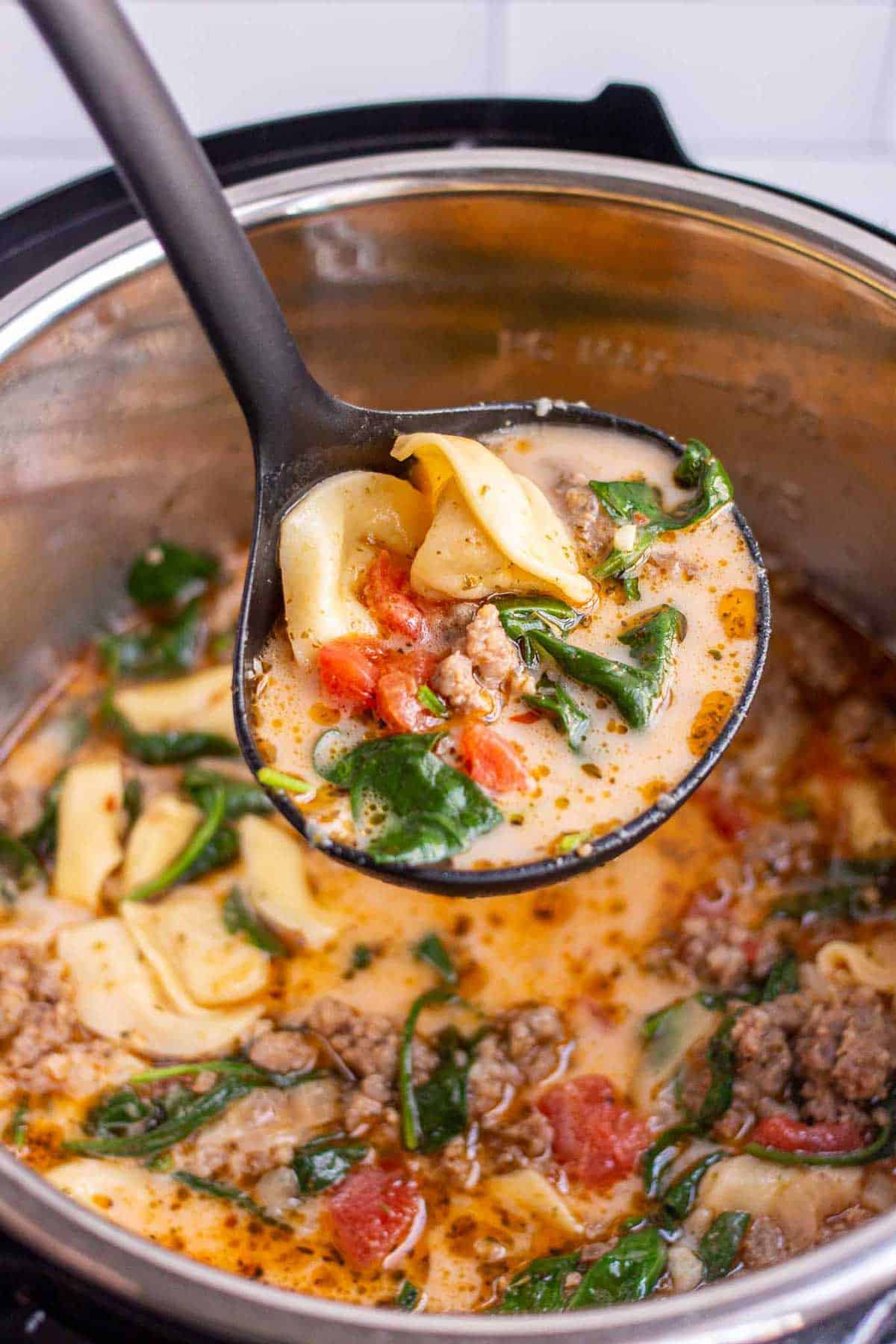Instant Pot Tortellini Soup with tortellini, Italian sausage, and wilted baby spinach in a ladle above the Instant Pot.