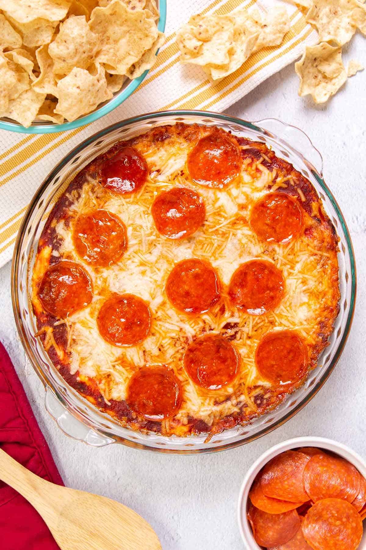 Top down view of pizza dip baked in a pie plate. The dip is topped with pepperoni. A bowl of tortilla chips is in the bowl off to the side.