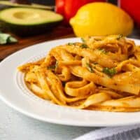 Avocado bell pepper pasta on a plate. Fettuccine noodles coated in sauce garnished with fresh basil. Avocado, red bell pepper, and lemon are in the background.