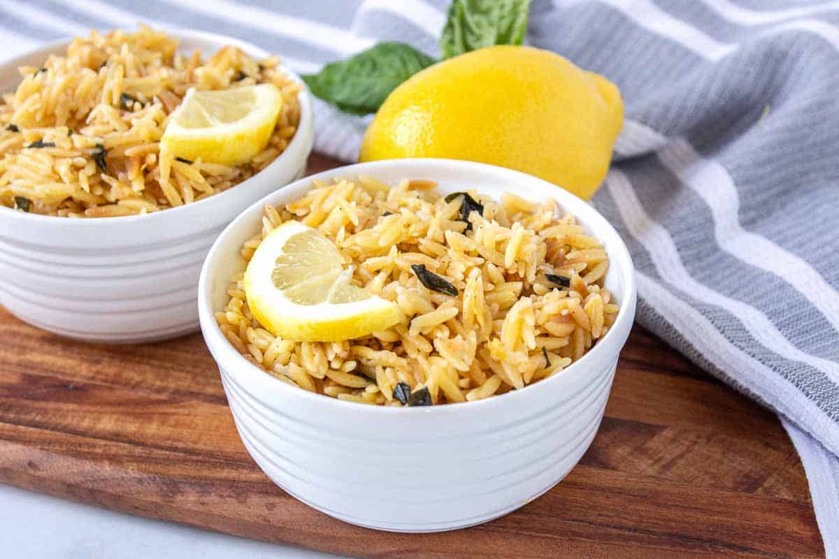 Ramekins filled with lemon parmesan orzo topped with a half lemon slice. There is fresh basil and lemon in the background.
