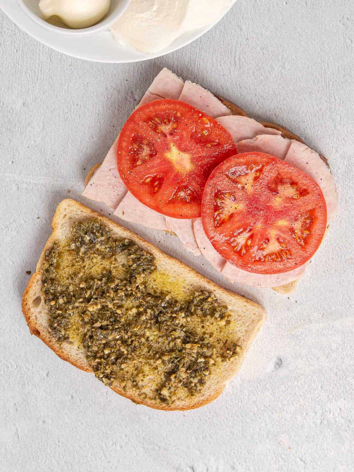 Two pieces of sourdough bread with pesto layered across the top. One one slice of bread, thick slices of turkey are layered on top with tomato slices on top of the turkey. The tomato slices have been salted and peppered.
