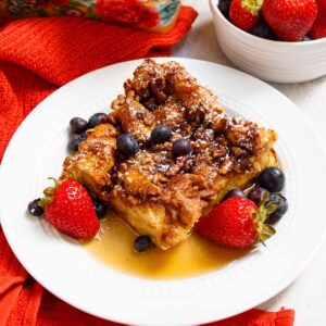 Square of French toast casserole served with berries, powdered sugar, and maple syrup.