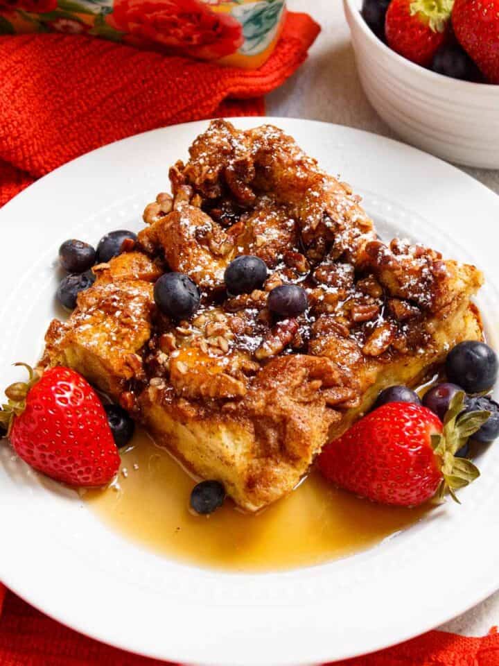 Square of French toast casserole served with berries, powdered sugar, and maple syrup.