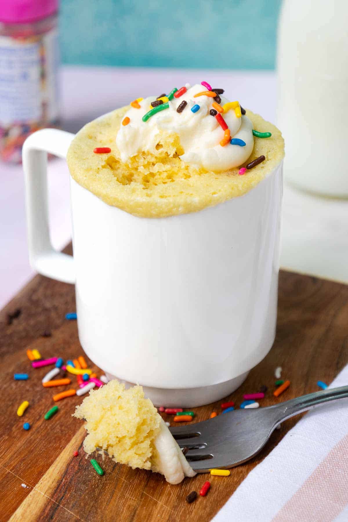 Yellow mug cake topped with buttercream frosting and sprinkles. A bite of cake rests on a fork to show the light texture.