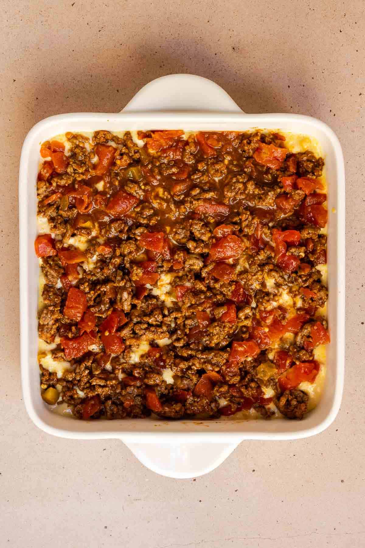 The ground beef mixture is spread evenly over top of the cornbread layer in the 8 by 8 inch baking dish.
