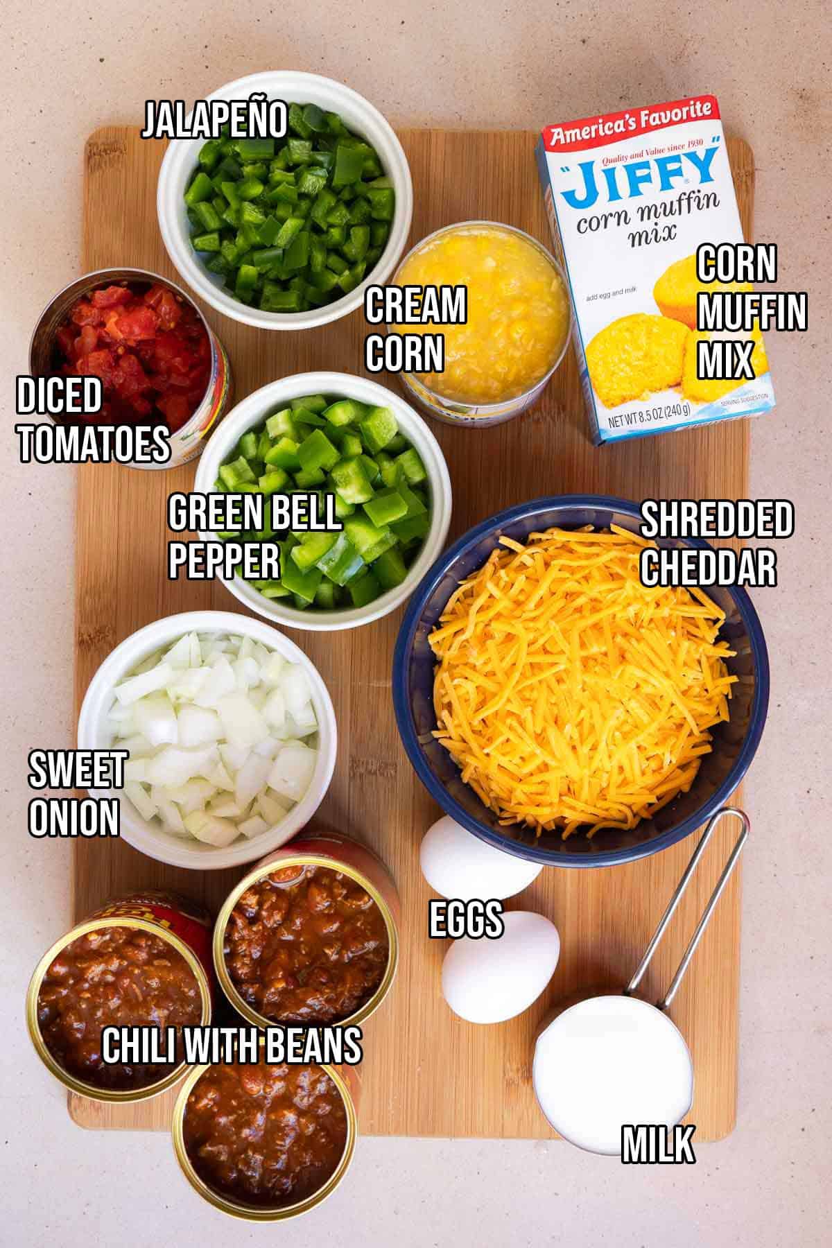 Chili cornbread casserole ingredients including canned chili with beans, fresh jalapeño, canned diced tomatoes, diced green bell pepper, diced sweet onion, shredded cheddar, a box of Jiffy corn muffin mix, canned cream style corn, eggs, and milk.