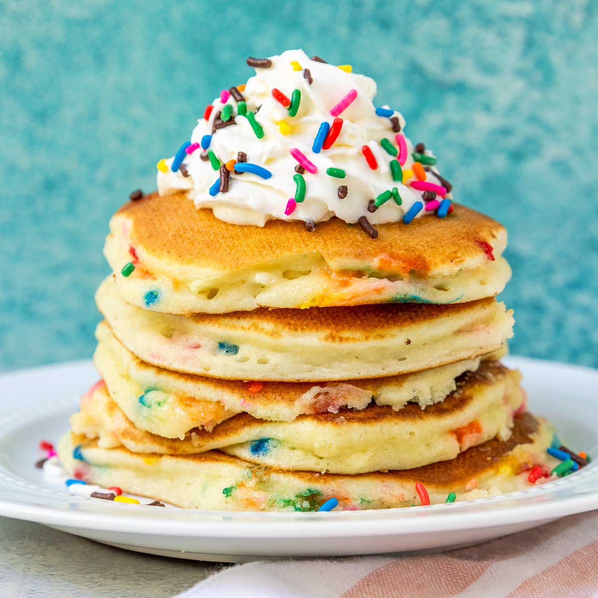 Stack of funfetti pancakes topped with whipped cream and rainbow sprinkles.