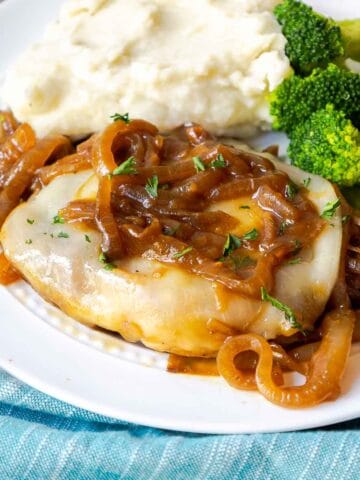 A pork chop topped with provolone and onions on a plate with mashed potatoes and broccoli.