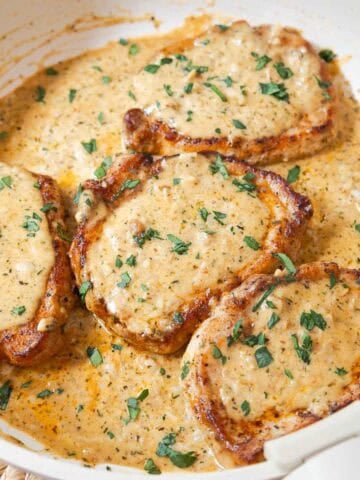 Four boneless pork chops in a skillet covered with creamy garlic sauce and fresh parsley.