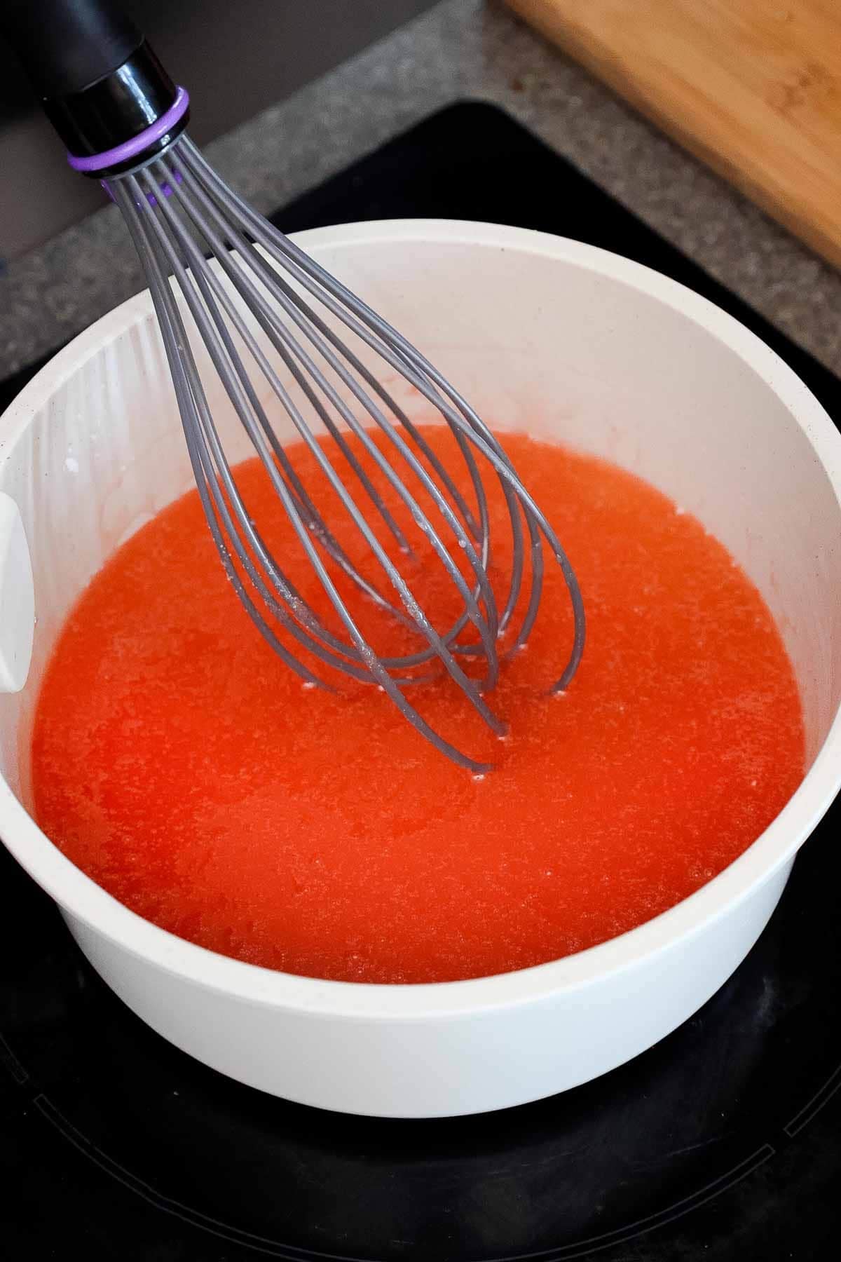 A saucepan with a whisk in a peach gelatin mixture.