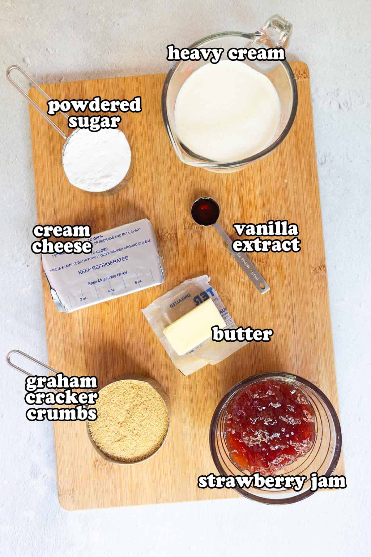 Ingredients on a wooden board: cream cheese, powdered sugar, heavy cream, vanilla extract, butter, graham cracker crumbs, and strawberry jam.