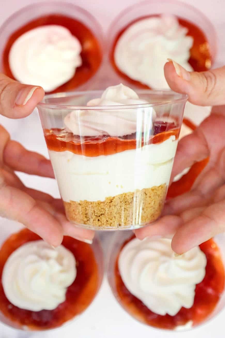 Hands holding a finished cheesecake cup topped with whipped cream. Other cups with whipped topping are in the background.