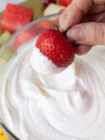 A hand holding a fresh strawberry dipped in fluffy marshmallow fruit dip.