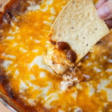 Tortilla chip scooping up hot chili cheese dip, revealing melted cheese, beans, and cream cheese.