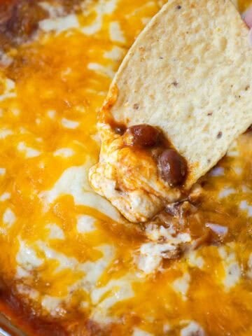 Tortilla chip scooping up hot chili cheese dip, revealing melted cheese, beans, and cream cheese.