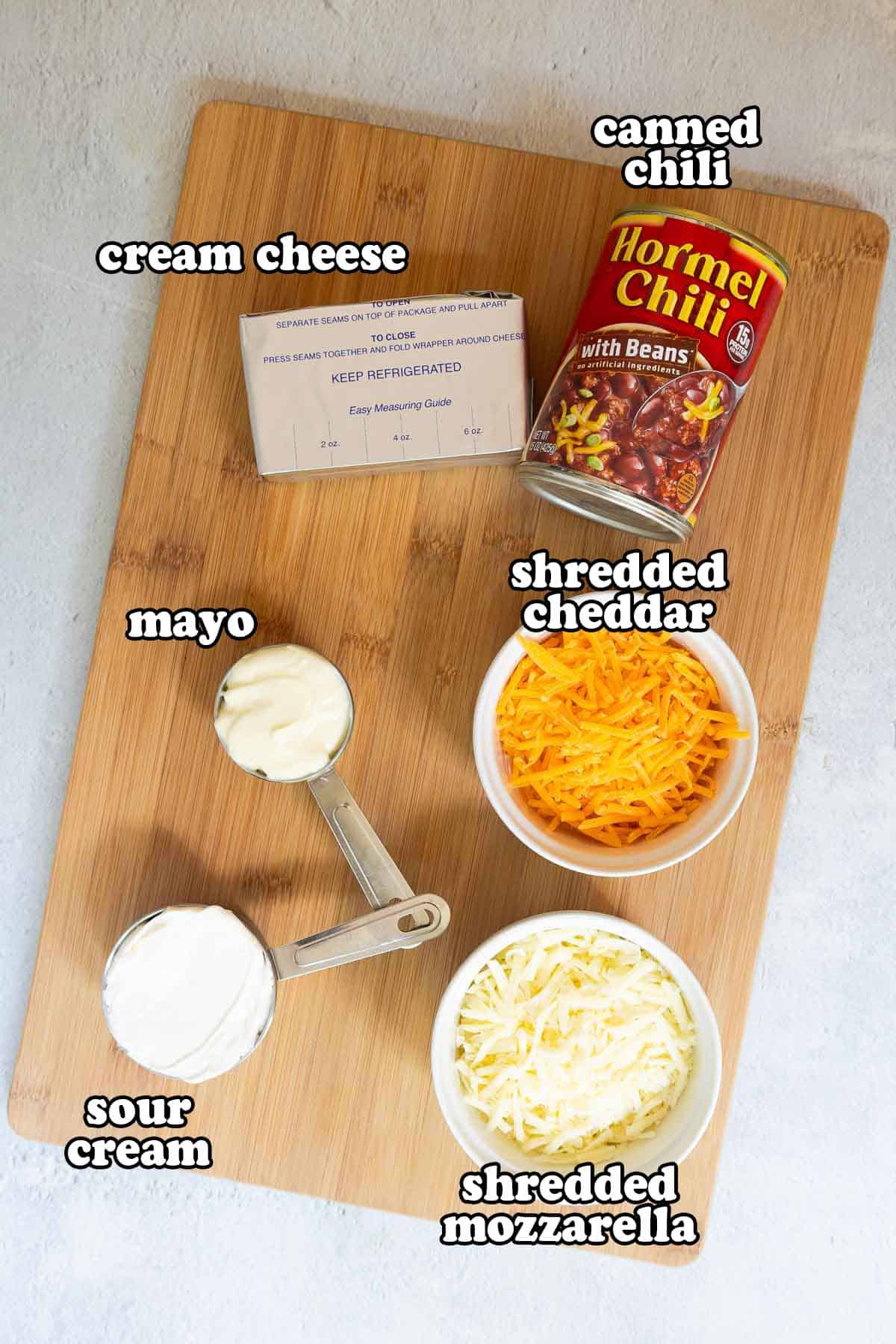 Overhead view of labeled chili cheese dip ingredients on a wooden cutting board, including canned chili, cream cheese, mayo, sour cream, shredded cheddar, and shredded mozzarella.