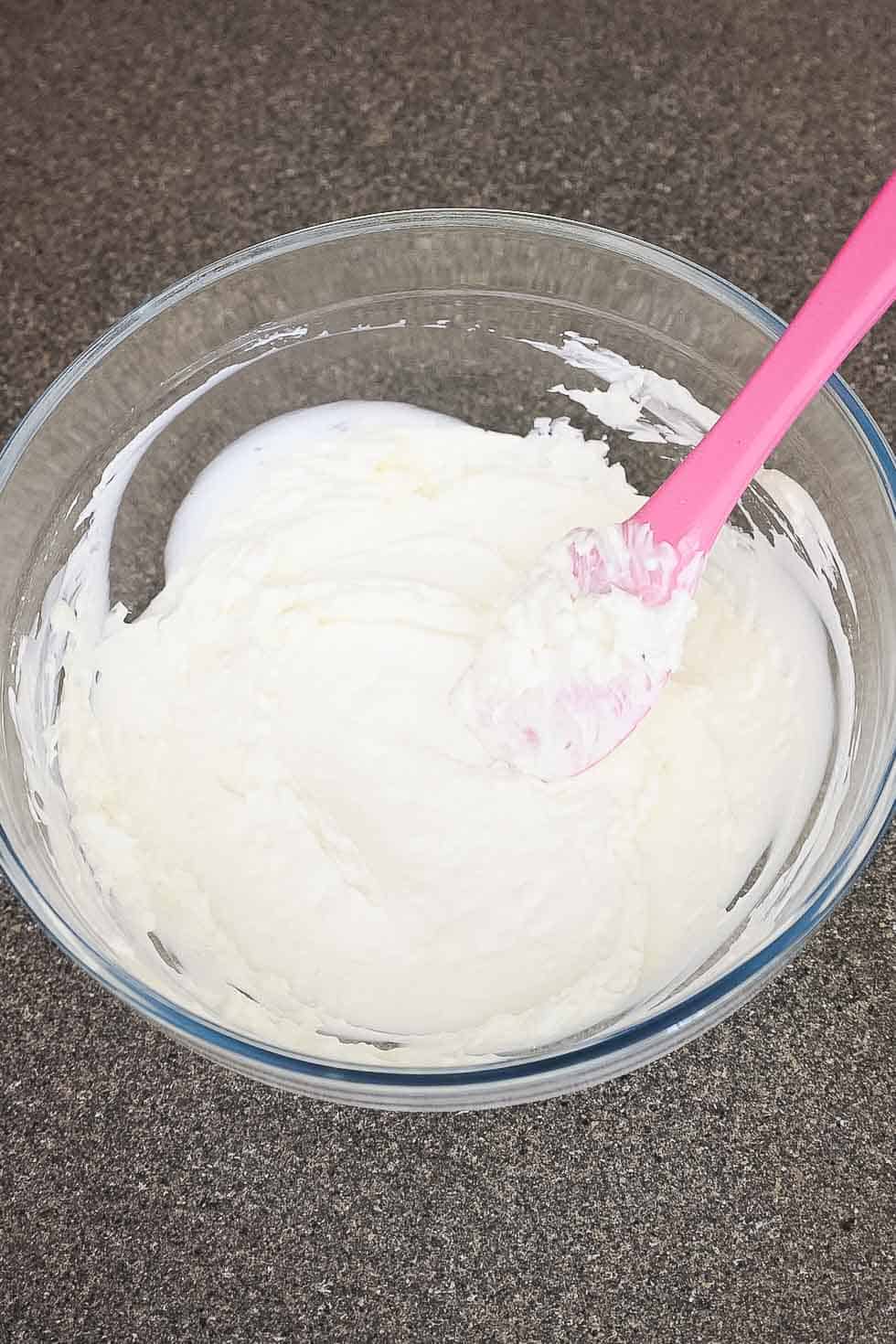 A glass bowl with a thick mixture of cream cheese, sour cream, and mayo stirred together with a pink spatula.