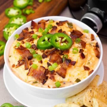 Small bowl of creamy crockpot jalapeño popper dip topped with crispy bacon, sliced jalapeños, and green onions.
