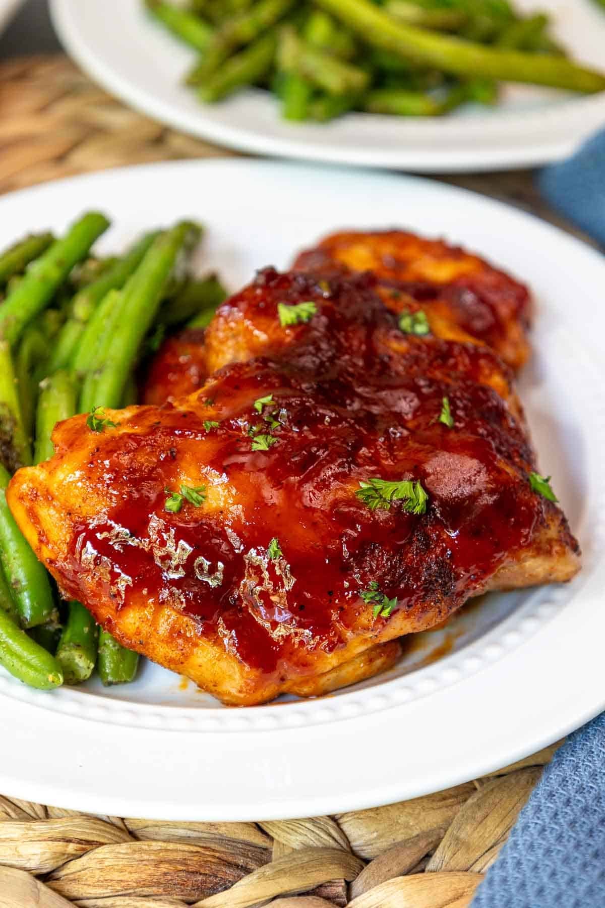 Close-up of oven baked BBQ boneless chicken thighs glazed with sauce and sprinkled with parsley, served with green beans on a white plate.