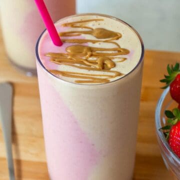A tall glass of peanut butter and jelly smoothie with pink and beige side-by-side in the glass, topped with peanut butter drizzle.