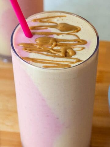 A tall glass of peanut butter and jelly smoothie with pink and beige side-by-side in the glass, topped with peanut butter drizzle.