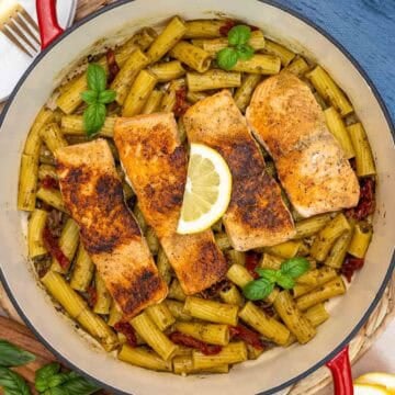 Pan of salmon pesto pasta with four seared salmon fillets on top of rigatoni tossed in pesto sauce, sun-dried tomatoes, and garnished with fresh basil and a lemon wedge.