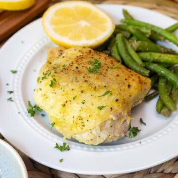 Close-up of a plated lemon pepper chicken thigh with crispy skin, paired with green beans and half a lemon for garnish.