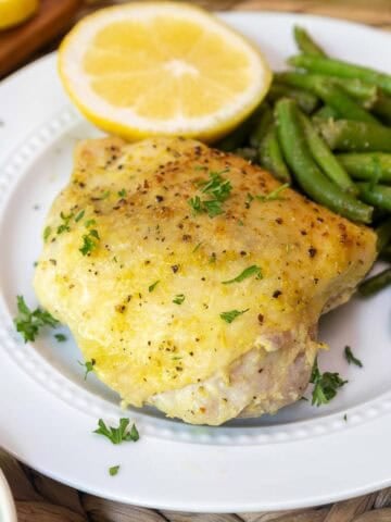 Close-up of a plated lemon pepper chicken thigh with crispy skin, paired with green beans and half a lemon for garnish.