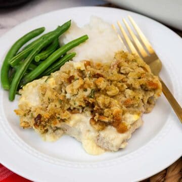 A serving of baked pork chops with stuffing, mashed potatoes, and green beans on a white plate with a gold fork.