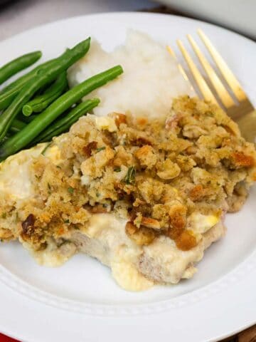A serving of baked pork chops with stuffing, mashed potatoes, and green beans on a white plate with a gold fork.